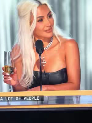 The cover image features Kim Kardashian, blonde hair in a half-updo, at a podium holding a glass of champagne. Her hair has a side part and is styled with loose waves framing her face. She wears a black strapless dress, a silver necklace, and a cross pendant. A microphone is positioned in front of her. Below, a banner reads '...' with the text out of frame.