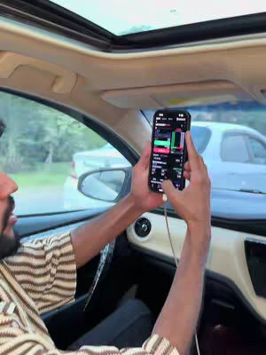 A man is seated inside a car, holding a smartphone displaying a trading application. The scene is shot from a first-person perspective, looking out through the windshield. The car's interior is visible, and the background shows a suburban road with trees and another car. The man appears to be in his early to mid-30s, wearing a patterned shirt. The phone screen dominates the shot, displaying charts and data relevant to stock trading. The overall composition gives a feeling of a work environment.