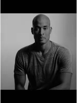 A black and white portrait of a bald man in a grey t-shirt against a light background. He has a serious expression and is looking directly at the camera. The lighting creates shadows, emphasizing his features and the texture of the clothing.