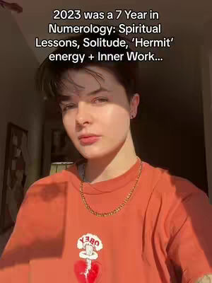 A person with short dark hair in an orange t-shirt, a gold chain necklace, and a red heart with an attached flower in front of their chest. The text overlay at the top provides the video's primary focus, referencing numerology and a specific year.