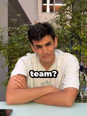 A young man with dark hair and a white t-shirt is seated at a table, arms crossed. A text overlay over the man’s torso asks “team?” The background includes natural greenery and a glimpse of a building.
