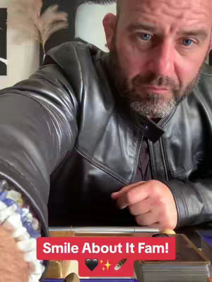 A man with a bald head and a short beard is the focus of the image, wearing a black leather jacket. Text overlay at the bottom states: "Smile About It Fam!" with emojis including a black heart. Part of a bracelet and a deck of cards are at the bottom