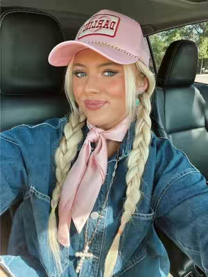 A young woman with long blonde braids sits in a car, wearing a pink baseball cap, denim jacket, and a pink scarf. She has a cross necklace. The image is well-lit, and she is looking at the camera.
