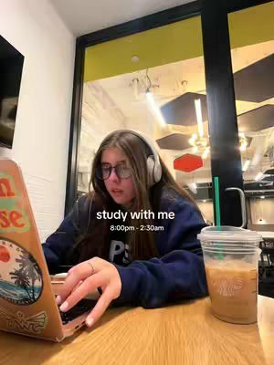 A young woman with long brown hair is seated at a wooden table in what appears to be a cafe or study area. She is wearing glasses and headphones, and has a laptop open in front of her. A coffee drink is next to the laptop. The background includes a large window and overhead lighting creating a lit study atmosphere.
