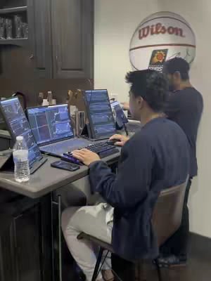 Two men are seated at a counter with several computer monitors displaying financial charts. A basketball with the words 'Wilson' is in the background. One man is facing away, focusing on his keyboard, while the other is partially visible, looking at the screens. The scene is lit with neutral lighting, and the overall composition suggests a workspace or study environment.