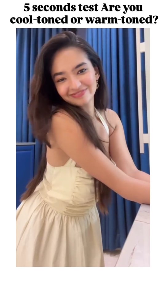 A young woman with long, dark hair smiles at the camera. She is wearing a light-colored dress and appears to be leaning against a table. The background includes blue drapes.