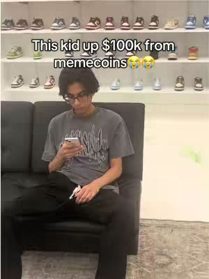 A young man with glasses sits on a black couch, looking at his phone. Behind him are shelves filled with sneakers. Text overlay reads, "This kid up $100k from memecoins" with crying laughing emojis.