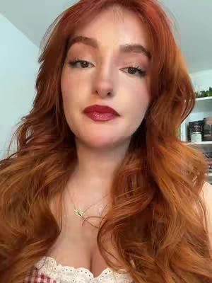 A woman with long, wavy red hair and red lipstick looks directly at the camera. She wears a delicate necklace and a white top. The background is blurred, with a hint of a white wall and a shelf with items.