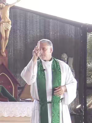 A medium shot shows a man in a white robe with a green sash, holding up a small piece of wood. Behind him is a crucifixion statue and another statue, and a dark, undefined background. The lighting is bright but soft, suggesting an outdoor setting.