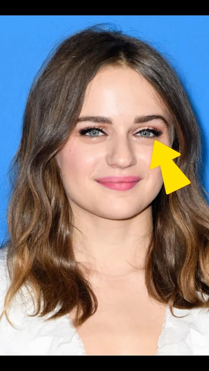The image is a close-up portrait of Joey King with a light blue background. She is looking at the camera with a slight smile and her eyes are highlighted with makeup emphasizing her eyes and a yellow arrow is pointing to the right eye. She has wavy brown hair, pink lipstick, and a white top.