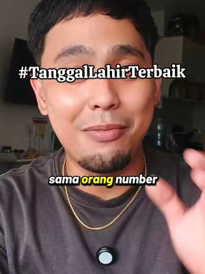 A man with dark hair and a short beard is centered in the frame. He's wearing a dark gray t-shirt and a gold chain. Overlaid on the image are two lines of white text: "#TanggalLahirTerbaik" at the top and "sama orang number" below. The background is blurred, suggesting an indoor setting.
