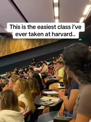 The image captures a classroom setting at Harvard University. The composition shows a lecture hall filled with students, most of whom are focused on the front. The lighting appears to be bright, likely from overhead fixtures, and the colors are muted. The layout of the room, with its tiered seating, suggests an academic environment. The overall composition is documentary, capturing an everyday scene.