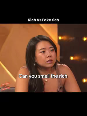 The cover image features a woman with dark hair in a medium shot. The top half of the image has a black background with the text "Rich Vs Fake rich" in white. The woman is centered and framed from the chest up, looking slightly to the left with a concerned expression. Text overlay "Can you smell the rich," appears to the bottom-center. Behind her is a blurred out background of what appears to be a stage setting, with warm colors.