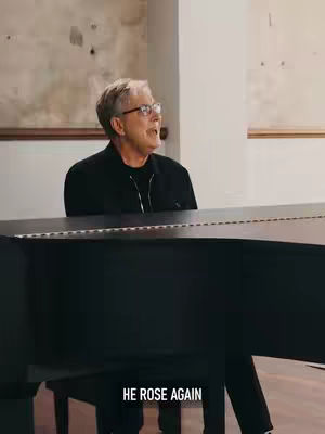 A man in glasses is seated at a piano, looking toward the right side of the frame. The piano is black, and the background shows a light-colored wall with some details, possibly in a church or hall.