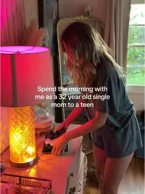 A woman is shown from the side, reaching for something on a white dresser. She's in a well-lit, cozy room with a large lamp and a mirror visible in the background. The image has a soft, warm lighting, with a pinkish-red lamp on the left and natural light from a window on the right.