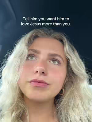 A selfie-style shot featuring a young woman looking upwards with a contemplative expression. The woman has long, wavy blonde hair and is wearing hoop earrings. She is positioned against a dark background, possibly inside a car. Text overlayed on the image reads: "Tell him you want him to love Jesus more than you."