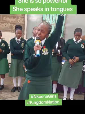 The image shows a group of young women in green and white uniforms, with one woman in the center holding a microphone and speaking. There are several other girls behind her. Text overlays are present, with phrases like 'She is so powerful' and '#NkueneGirls, #KingdomNation' visible.