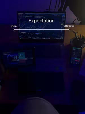 A dimly lit desk scene with multiple screens displaying what appears to be stock market data. The primary screen has text overlayed. A white line with circles at each end spans the screen, labeled "idea" on the left and "success" on the right, with "expectation" written above. There's a smartphone and another screen visible, all in dark, muted tones, hinting at a focused work environment.