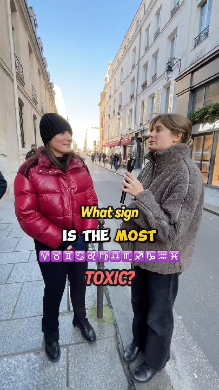 Two women are standing on a city street, possibly in Europe. One woman holds a microphone and appears to be interviewing the other. A text overlay asks “What sign IS THE MOST TOXIC?” with zodiac symbols below the question. The background includes buildings and a clear daytime sky.