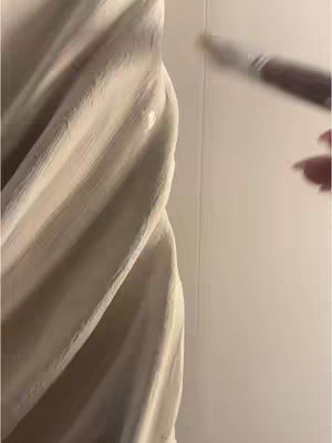 The image shows abstract and close-up detail of a piece of fabric. The fabric is a light, creamy color and appears to be draped or folded, with subtle textures and shadows hinting at its form. A hand holding a brush appears in the right part. The overall effect is minimalist and tactile, with soft lighting.