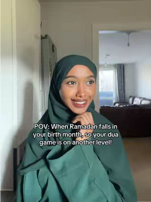A woman wearing a green hijab and top is smiling at the camera. Text overlayed on top of the video says, “POV: When Ramadan falls in your birth month, so your dua game is on another level!”. The background of the video is a home interior with a white wall on the left side and a doorway on the right side which opens to another room, with a window.