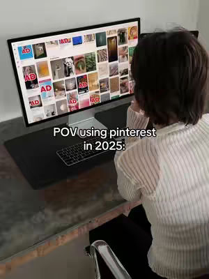 The image shows a woman sitting at a desk, facing a large computer monitor. The monitor displays a Pinterest-like interface, filled with various images. The text overlay reads, 'POV using pinterest in 2025:'. The woman is seen from the back, with short dark hair, wearing a white sweater, looking at the screen which displays content labeled with 'AD'. The workspace includes a keyboard and a black desk pad, it has a calm and focused atmosphere.