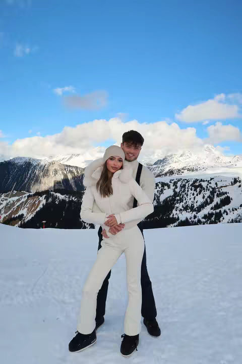 A couple stands in a snowy mountain landscape, with a bright blue sky overhead. The woman is in the foreground wearing a white ski suit and a fur collar. The man is behind her, also in white, with darker pants, his arms wrapped around her. The snow-covered mountains create a scenic backdrop.