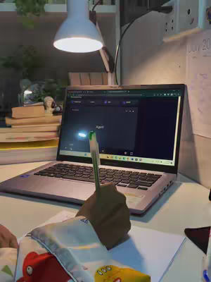 A well-lit workspace featuring a laptop, pen, and notes, suggesting a study session. A bright desk lamp illuminates the scene, and a hand holding the pen is visible in the foreground. The laptop screen displays what looks like a video call.