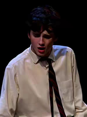 The cover image features a young man, likely Timothée Chalamet, in what appears to be a theatrical setting. He is facing slightly downwards, his mouth open as if speaking or singing, and has a focused expression. He wears a light-colored collared shirt, a dark tie with stripes, and his dark hair is styled with some volume. The background is a solid dark color, which emphasizes the man in the foreground. The lighting appears to be focused on the performer.