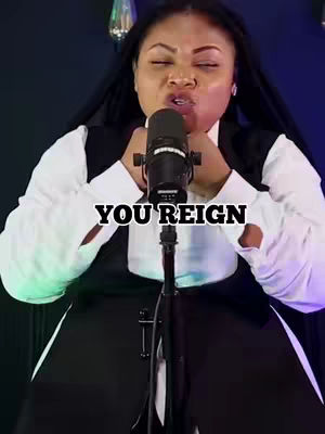 A medium shot captures a woman in a white blouse and black vest, passionately singing into a black microphone. Overlaid on the image is the text 'YOU REIGN,' centered below the microphone. The background is blurred with lights.