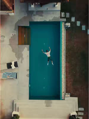 An aerial shot shows a rectangular swimming pool filled with turquoise water. A man in a t-shirt and shorts is floating in the center of the pool with his arms outstretched, as if sunbathing or looking up. The pool is bordered by a concrete deck with lounge chairs and a striped towel. The surrounding area includes a building with an orange door and a grassy area with stone stepping stones.