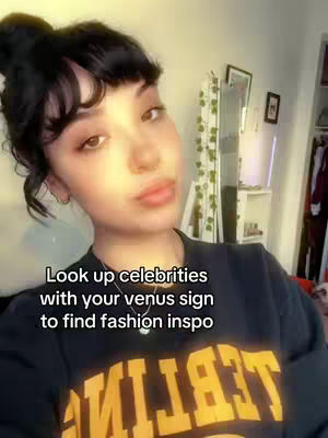 A young woman with dark hair styled in a bun and bangs is looking towards the camera, with text overlayed on her chest. She has light skin and is wearing a dark t-shirt with yellow lettering. The background is slightly blurred and shows hints of a room with wall decorations. The central focus is on the woman.