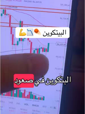 A screenshot of a phone screen displaying a stock chart. The chart shows a mix of green and red candlesticks, indicating market fluctuations. Overlayed on the screen are text boxes highlighting the word 'Bitcoin' in Arabic. A finger is partially visible, pointing toward the screen, and there are also two text boxes in the image, one at the top and one at the bottom. The background of the image appears to be a blurry mix of colors, likely from the surrounding environment. Overall, the composition presents a straightforward visual presentation of bitcoin.