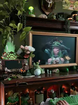 A cozy domestic setup featuring a wooden cabinet filled with various green objects and plants, creating a warm and inviting atmosphere. A framed artwork on a TV screen displays a humanoid figure with a floral headdress. The scene is filled with a variety of decorative elements, including glowing accents and a plush toy.