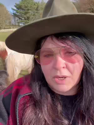 A woman wearing a fedora, sunglasses, and a plaid shirt looks directly at the camera, with a horse and trees in the background.