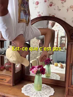 The cover image shows a person hanging upside down in a room with a mirror. The person is the main focus, with the reflection in the mirror showing a more complete view of the room. The room itself appears to be a vintage setting with wooden furniture, a floral wallpaper, and a vase of flowers. The text overlay reads 'Est-ce que c'est pas'.