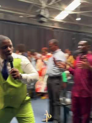 The cover image shows a medium shot of what appears to be a church service. A man in a green vest and white long sleeve shirt is holding a microphone and speaking. Behind him, other individuals are visible, and the background is blurred, indicating a large gathering. The lighting suggests an indoor setting.
