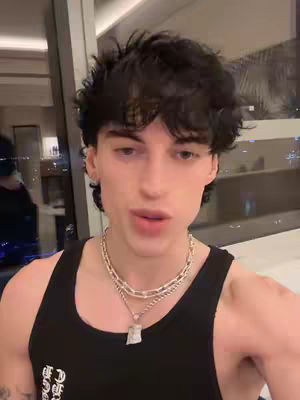 A young man with dark, tousled hair and a focused expression is the main subject. He's wearing a black tank top and multiple silver chain necklaces. The background is blurred, suggesting an indoor setting with a window showing city lights.