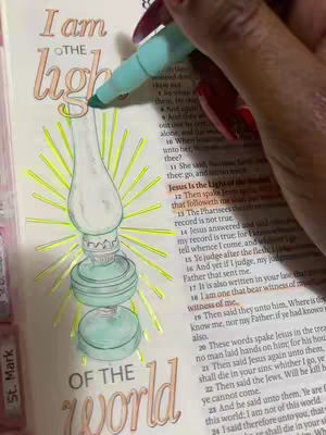 A person's hand with red nail polish holding a teal marker, coloring the illustration of a lamp in a Bible. The lamp is surrounded by yellow rays. The words 'I am the light of the world' are visible.