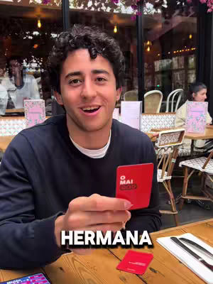 A young man with curly dark hair and a navy sweater smiles. He is sitting at a wooden table, holding a red card that says 'O Mai' on it, and there's a smaller red card and some phone in front of him. The word 'HERMANA' is overlaid across the bottom of the image.