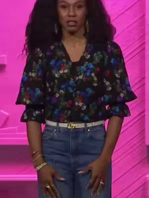 A woman with long, dark, curly hair stands against a light pink background. She wears a black blouse with a floral pattern in blue, red, and white, with flared sleeves, paired with blue jeans and a white belt with a gold buckle. She also wears bracelets and rings. She is looking directly at the camera with a serious expression.