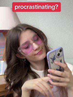 A young woman with long brown hair wearing large, pink-tinted square glasses holds a smartphone in front of her. The phone has a butterfly case. She is looking down at the phone with a slight smile. A red banner above her head reads, "procrastinating?" The background shows a lamp and a wall.