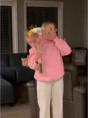 A woman wearing a pink hoodie and white pants is in a room, holding a bouquet of flowers and covering her mouth with her hand, with a surprised expression. The background is blurred, with a couch and windows with blinds visible.