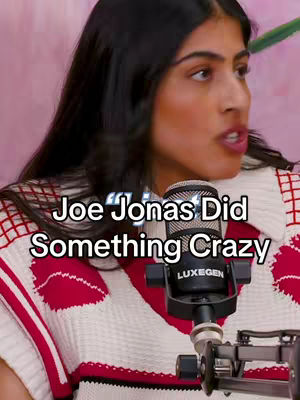 A woman with dark hair is speaking into a microphone. Text overlayed on her face says 'Joe Jonas Did Something Crazy'. The background is a soft, blurred pink and shows parts of a white and red top.