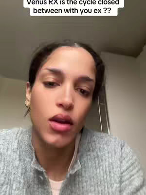 A close-up shot of a woman with dark hair and neutral makeup looking directly into the camera. She is wearing a gray sweater. In the background, a dim and blurry room with a few visible objects, including a lamp