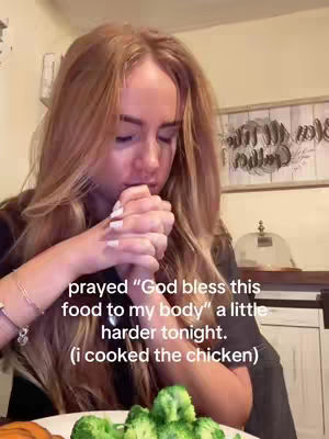 A woman with long blonde hair is shown from the chest up, with her head bowed and hands clasped together in prayer. She is seated at a table with a plate of food, including broccoli. The background shows a kitchen setting with a decorative wooden piece.