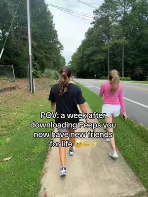 Two young women are captured from behind, walking on a sidewalk. They're casually dressed, with one in a black t-shirt and the other in a pink long-sleeve shirt. The setting appears to be a lush, green area with trees and a road in the background. The sky is overcast.