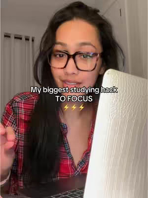 A young woman with dark hair and glasses looks directly at the camera, leaning slightly towards an open laptop on her right. She’s wearing a red and black plaid shirt. The background is a softly lit room with vertical blinds visible. Text overlays indicate the video's content.