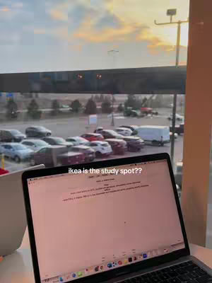 A well-lit laptop sits open on a table, with the screen displaying text. Through a large window, a parking lot filled with cars and a slightly cloudy sunset are visible. A sign reading "ikea is the study spot??" is superimposed on the image.