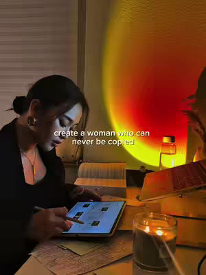 A dimly lit scene shows a woman studying at a desk. She is the primary focus, with a tablet in front of her and a pen in hand. A glowing orange light casts a warm hue and the text overlay reads: create a woman who can never be copied.